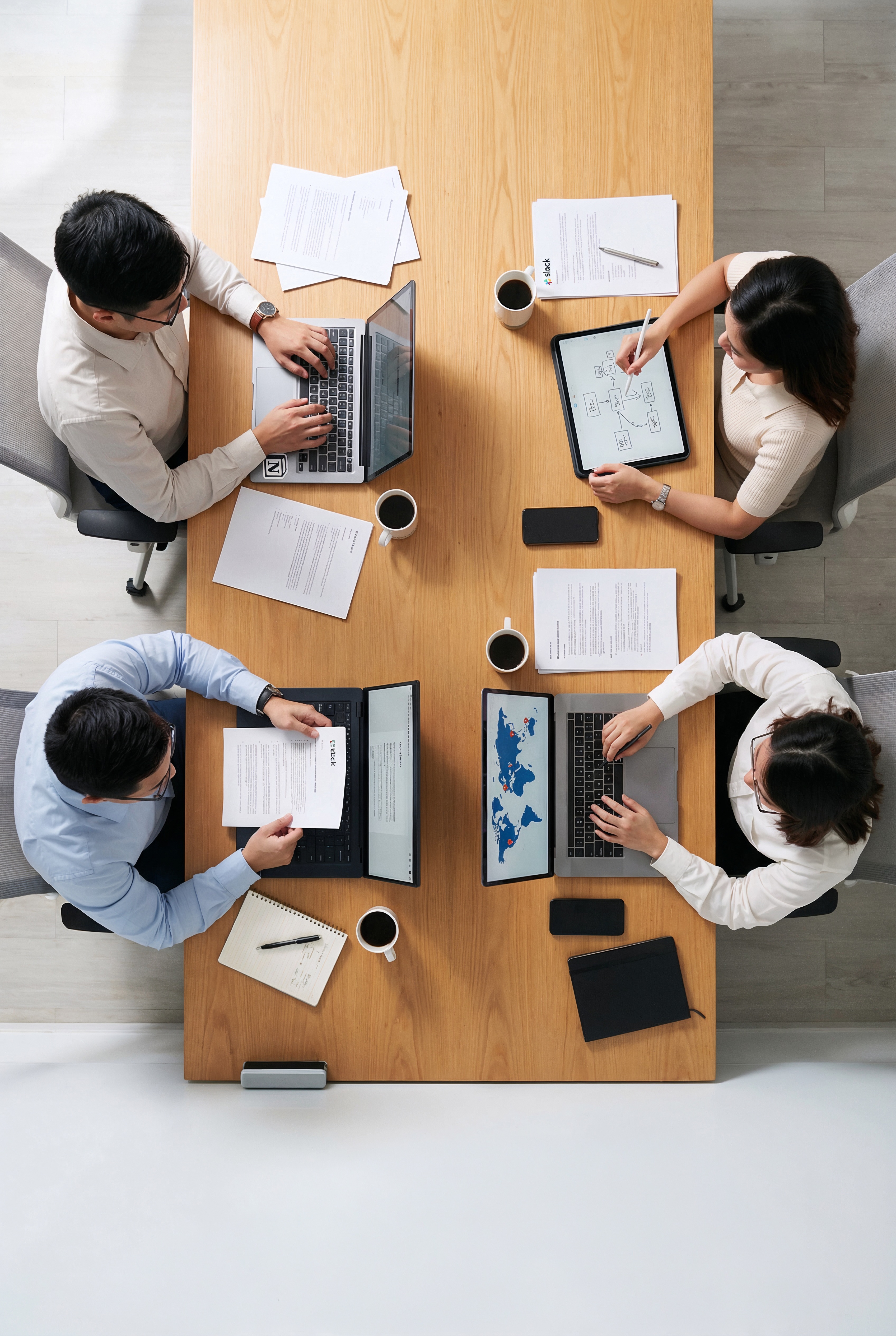 Four colleagues collaborating around a table, seen from above, working on laptops and tablets during a meeting.